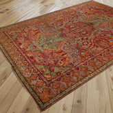 A traditional oriental area rug with an intricate floral and medallion pattern in soft pinkish red, marmalade, soft green, and touches of crushed raspberry, displayed on a light-coloured wooden plank floor.
