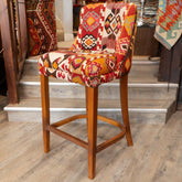 A wooden bar stool with a colorful geometric kilim patterned seat and backrest, set in a home interior with wooden stairs and rugs in the background.