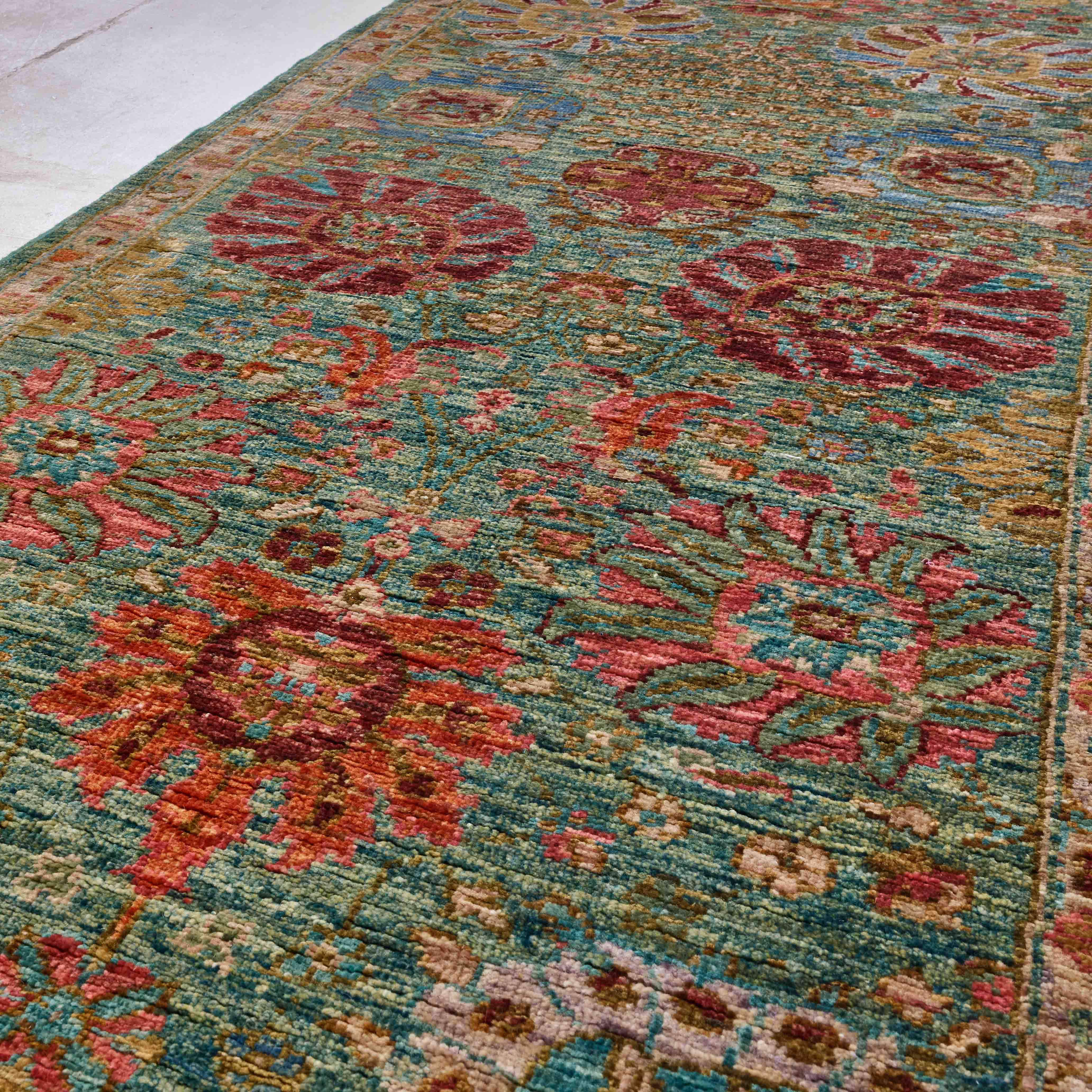 A close-up view of a traditional patterned runner with a teal blue-green base and large floral motifs in papaya spice, hibiscus pink, crimson, and muted gold.