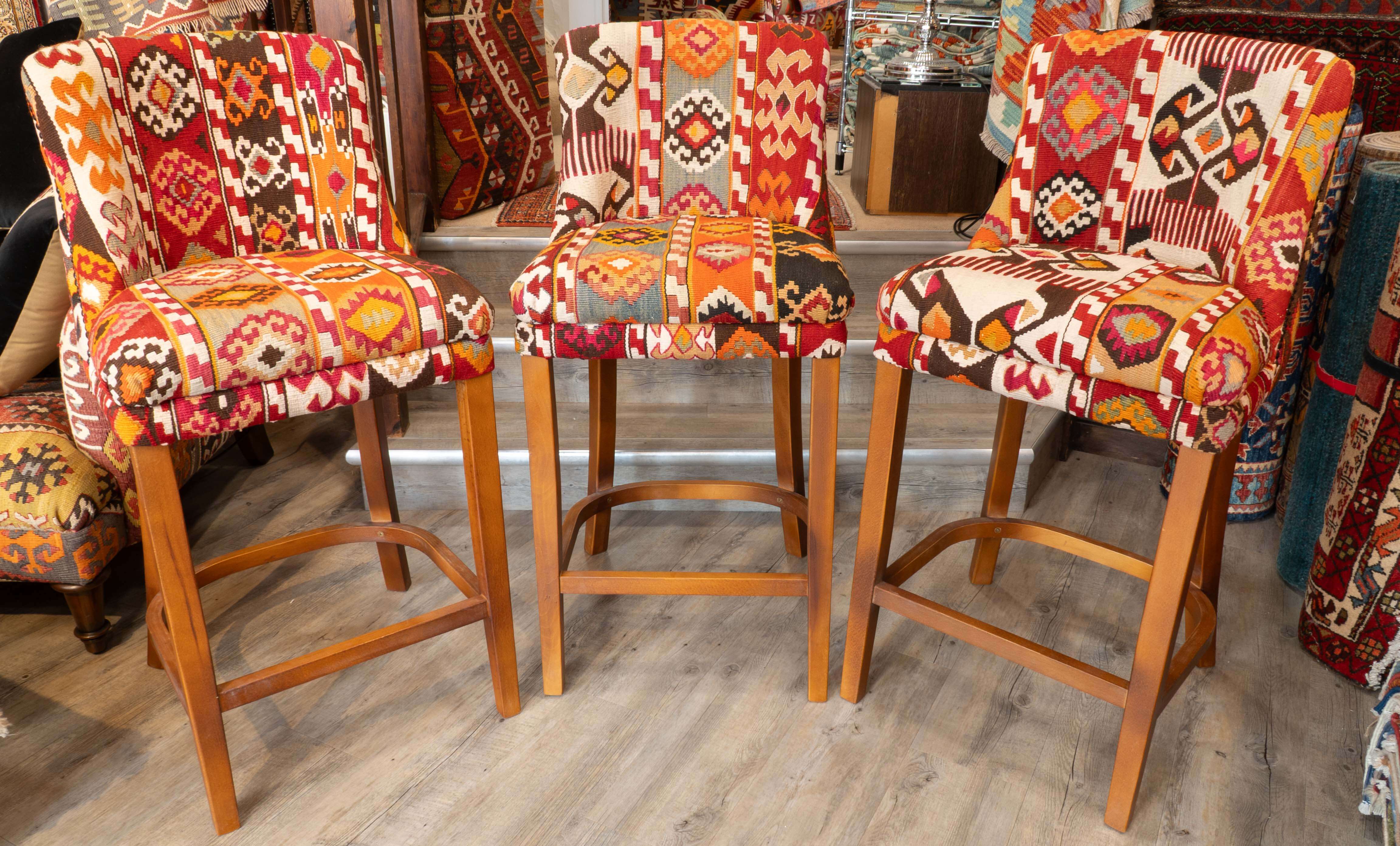 Three wooden bar stools with colourful geometric patterned upholstery in red, orange, beige, and black, displayed on a wood-look floor in a Jennings rug store.