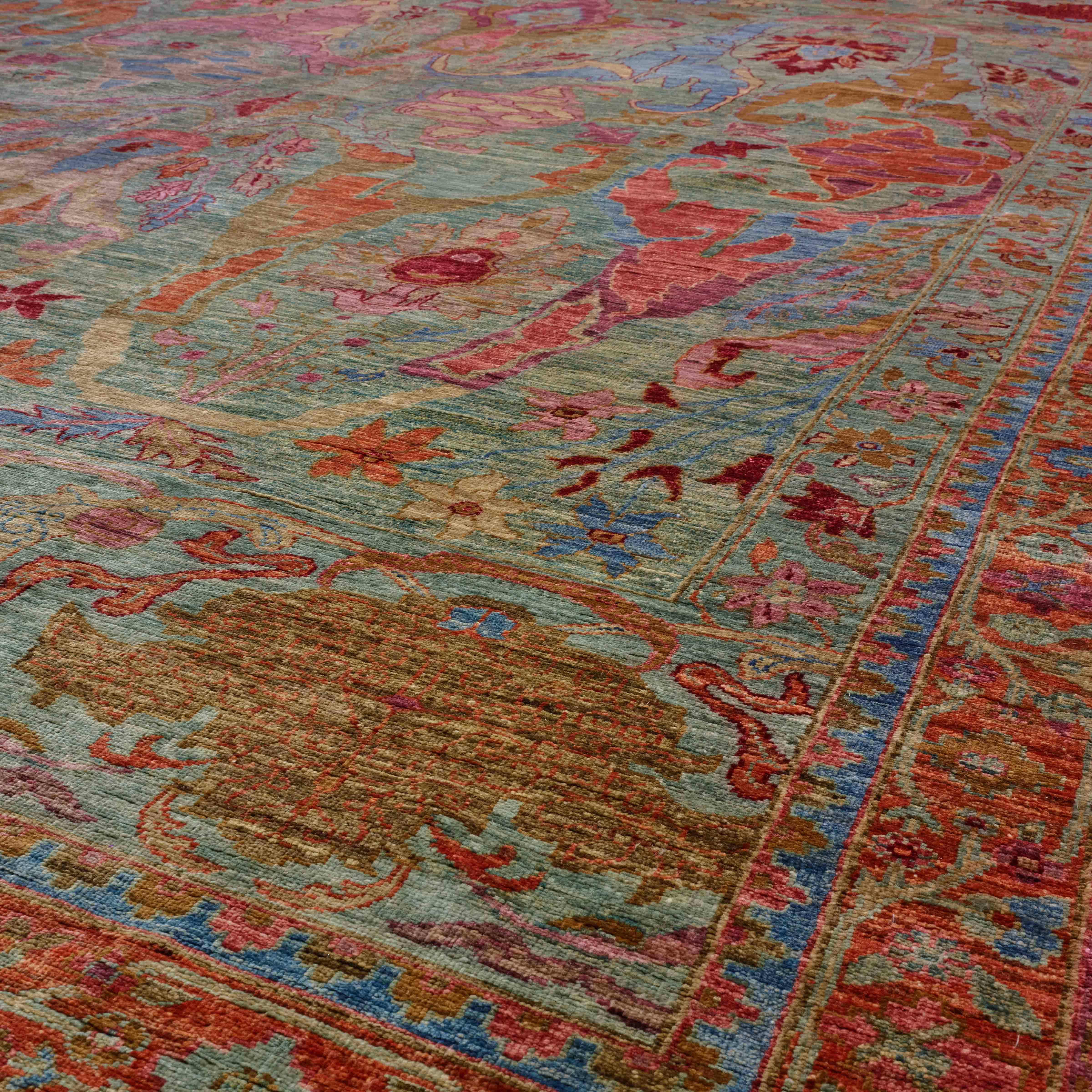 A close-up view of an ornate traditional area rug with a complex floral and medallion pattern in muted tones of sage green, terracotta red, dusty blue, and golden brown.