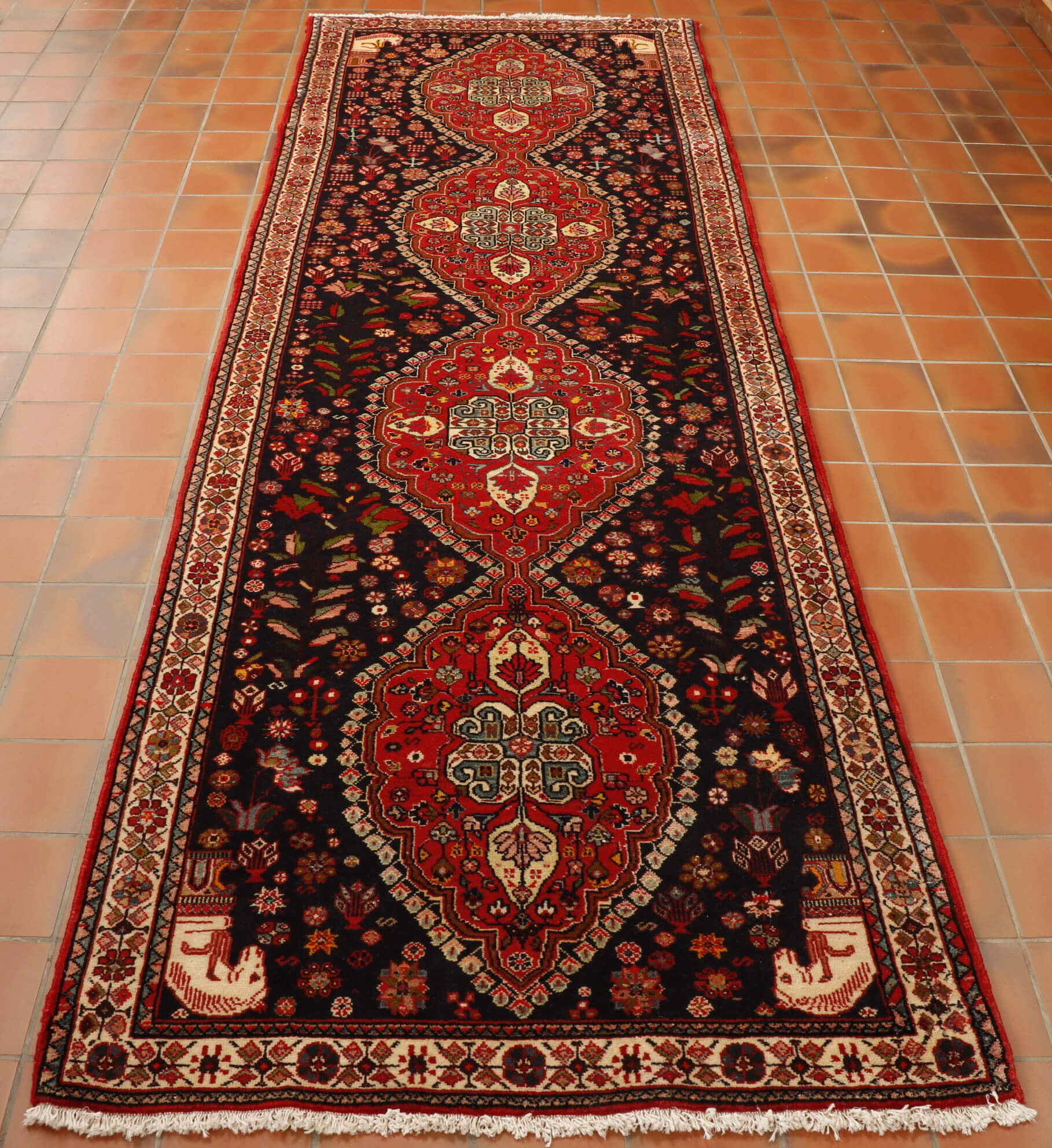 A deep blue ground holds 4 connected leaf shapes in the main area going through the length, these are in broght red.  There is a border in cream. In each corner there is a horses head in cream.  All over, all areas of this rug are depictions of flowers, birds and butterflies. 