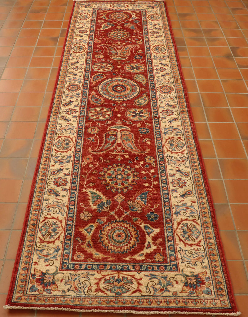 Russet red central ground with flora and fauna decoration through the length.  There is a central motif which is circular,  and two further circular motifs at either end.   The colours used within the colour palette for the decoration are green, blue, yellow, cream, and a soft orange.  There is a broad cream border with floral decoration again using the same colours.   
