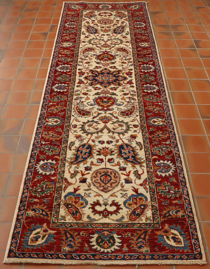 A striking Aryana runner with a cream central ground with flora and fauna design using greens, blues orange, red, peach and golden yellow.  There is a broad red border with contrasting design using colours from the same colour pallet. 