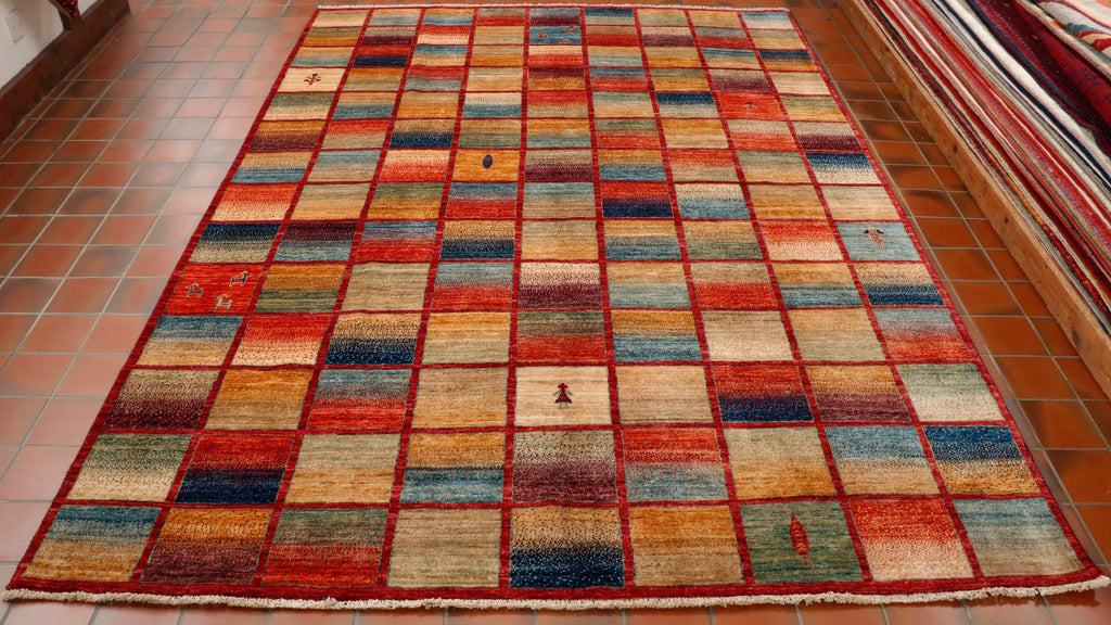 This piece is made up of a grid of colours.  9 across the width and 14 the length, in total therefore 126 boxes.   Each row on the grid has a red border breaking up the boxes.   The colour palette used in this rug are a range of blues, greens, yellows, oranges and plum.  Each box has two of the colours blended.  In a couple of the boxes there are small depictions of fauna, animals or humans.  