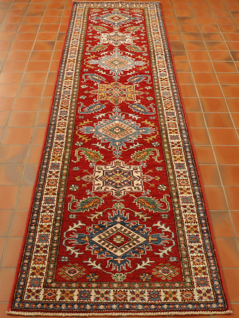 Strong red ground in the central section with 6 decorated motifs, laid out symmetrically through the length of the runner.  There are 3 main borders one being cream that is slightly broader than the other two.  All of the decoration has a colour palette of cream, blue, green, and yellow.  These vary in shade. The decorative featues are all based on stylized flora and fauna. 