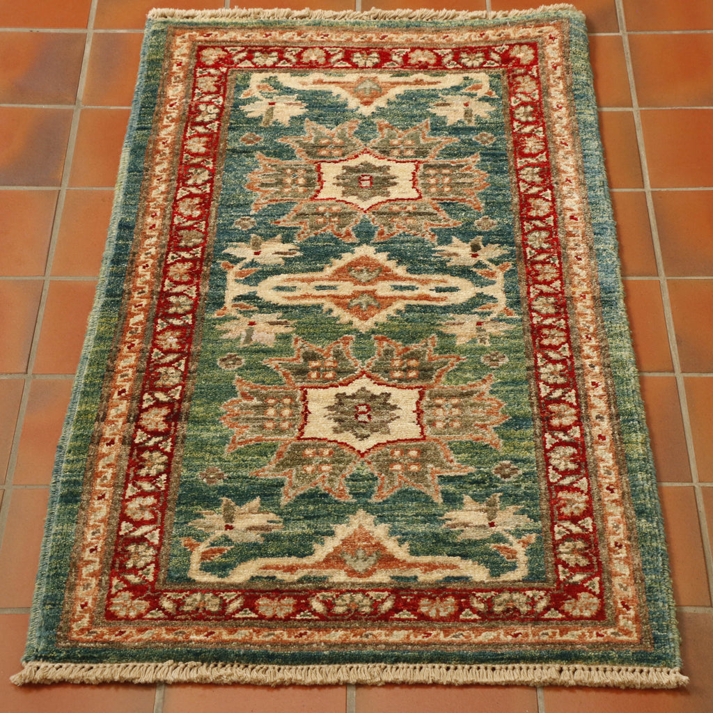 This rug has a greeny blue background, there are parts which are more green than blue. The decoration is stylized flowers.  A main one in the centre, two larger, different ones follow either side and then two half flowers, mirroring the design of the central one at each end.  All of these have a predominantly cream and terracotta palette.  