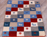 This small mat has a chequerboard design in various shades of blue, red, cream, grey and oatmeal.  There are small depictions in some of the sections of goats and stylized leaves using the same colour palette but contrasting.  